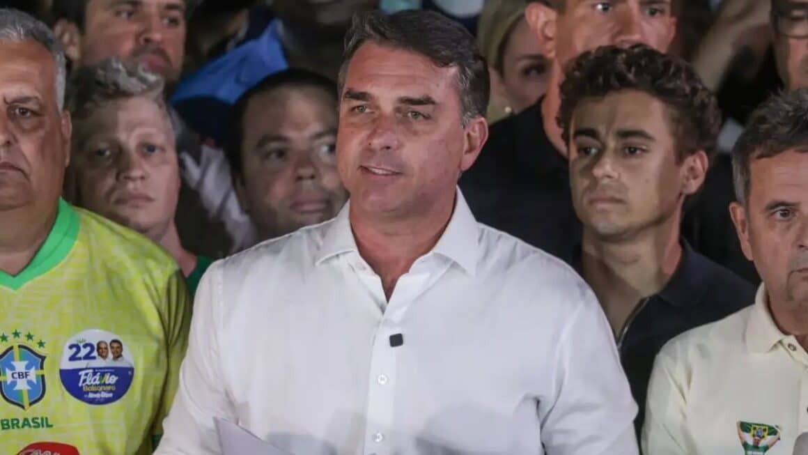 Flávio Bolsonaro deixa o Brasil e segue para os EUA, dizem aliados O senador Flávio Bolsonaro de camisa social branca discursa ao centro, cercado por apoiadores e aliados políticos ao seu redor em um pronunciamento público.