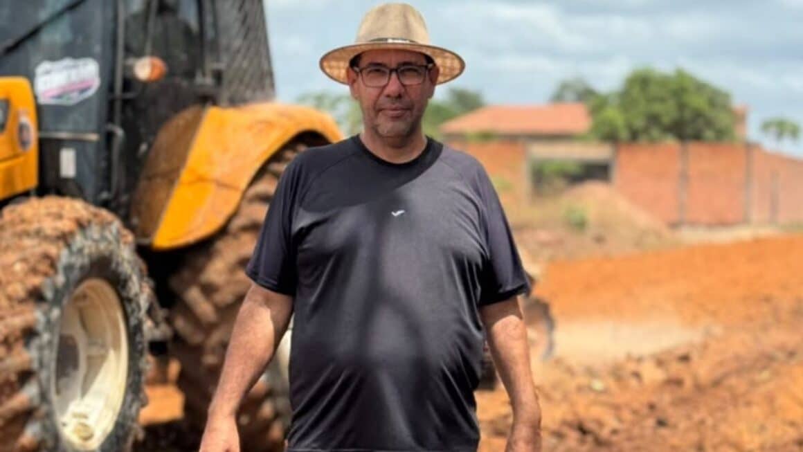 Demanda atendida: Miltinho Boiadeiro acompanha terraplanagem no bairro Fluminense O vereador Miltinho Boiadeiro de chapéu de palha, óculos e camiseta preta, em pé em frente a uma máquina amarela de terraplanagem na 7ª Avenida, em Tailândia.