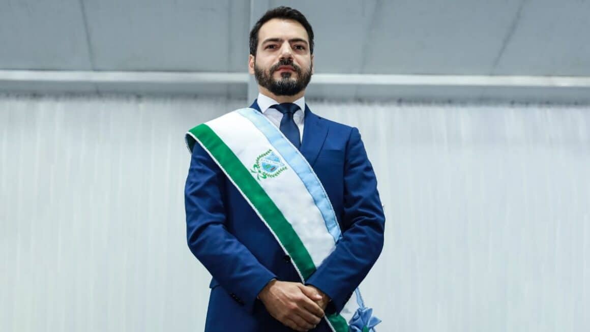 Qual o retrato da gestão de Eduardo Pio X em São Miguel do Guamá? O prefeito Eduardo Pio X de terno azul e faixa governamental transversal verde, branca e azul-claro posa em pé com as mãos cruzadas à frente em cerimônia oficial.