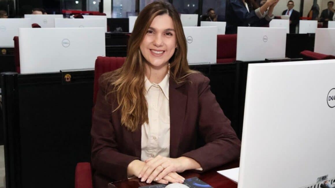 Deputada Paula Titan: trabalho parlamentar e novas conexões pelo Pará A deputada estadual Paula Titan sorri sentada em sua bancada no plenário da Assembleia Legislativa do Pará, vestindo blazer marrom-café sobre camisa clara.