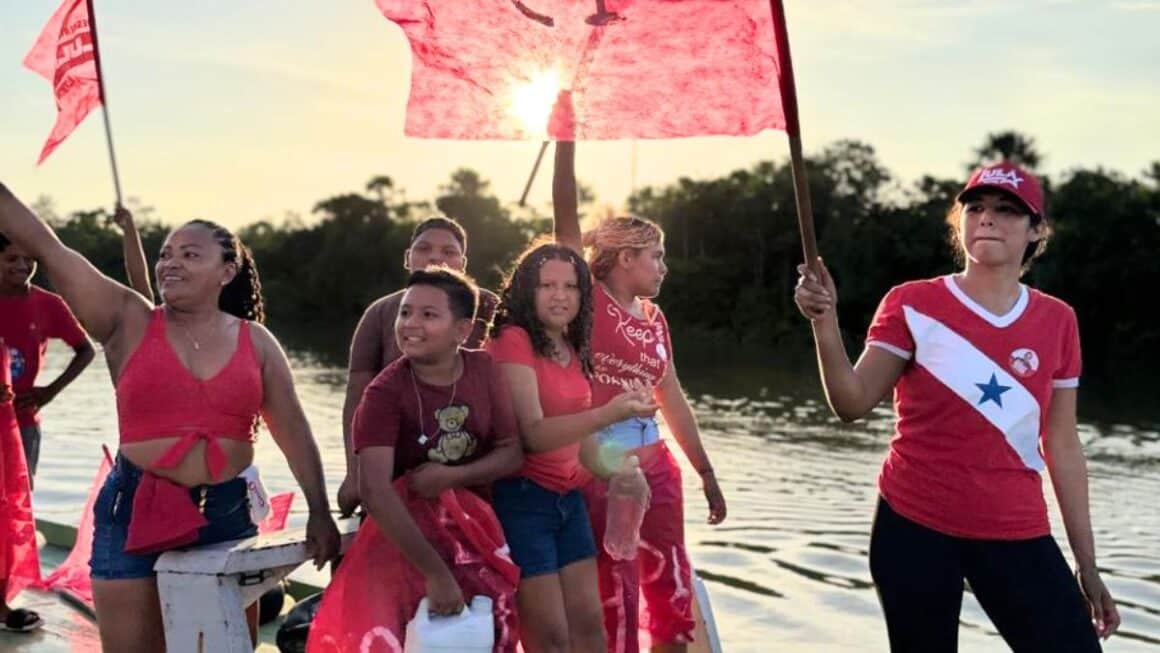 Jovem ribeirinha e quilombola, Ana Cláudia Neri, das Ilhas de Abaetetuba ganhou destaque na campanha da reeleição do presidente Lula no município Grupo de moradores e militantes ribeirinhos navegando de barco pelos rios das Ilhas de Abaetetuba, segurando bandeiras vermelhas ao por do sol, com a jovem liderança Ana Cláudia Neri em destaque à direita de camisa vermelha com estrela branca e boné.