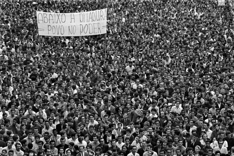 Fotografia aérea a preto e branco mostrando uma massa compacta e densa de manifestantes, na sua maioria jovens, preenchendo completamente uma avenida. Acima da multidão, um cartaz manuscrito e bem visível lê-se: "ABAIXO A DITADURA POVO NO PODER".