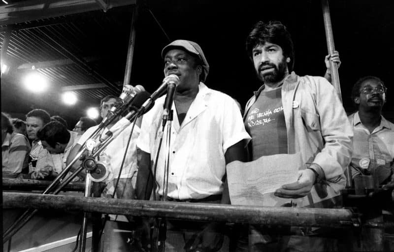 Fotografia a preto e branco de Milton Nascimento a falar ao microfone num palanque, acompanhado por outro homem que segura papéis durante um evento público de mobilização.